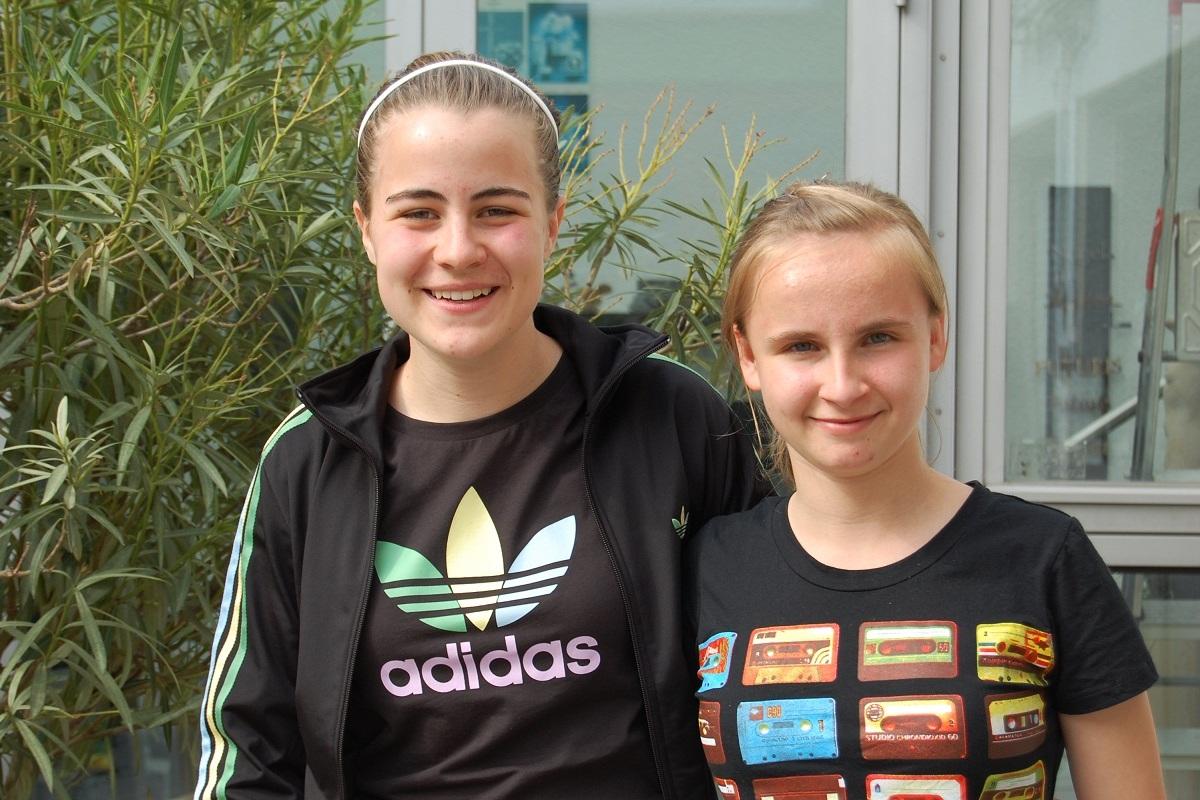 Zwei junge Frauen stehen lächelnd vor einer Pflanze, die Fenster eines Gebäudes im Hintergrund.