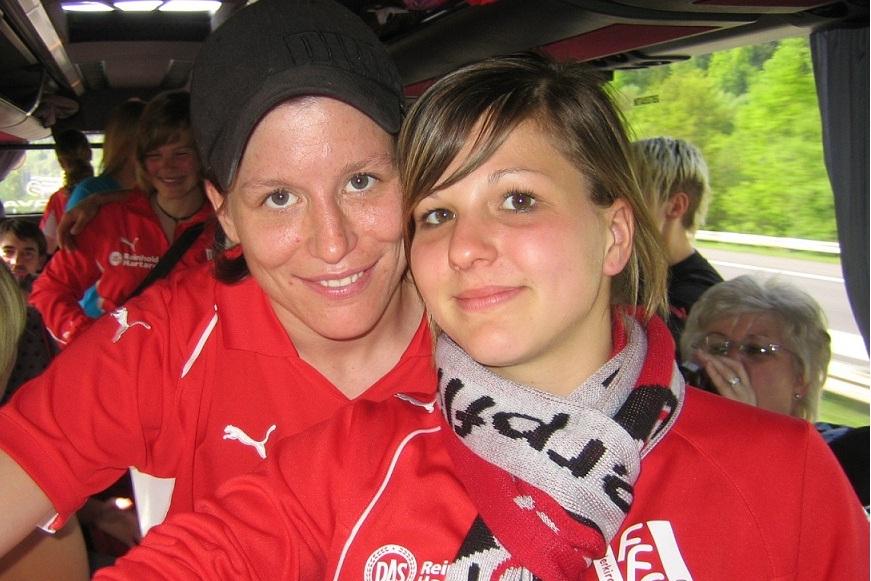 Zwei junge Frauen mit Fu&szlig;balltrikots und Schals l&auml;cheln in einem Reisebus, im Hintergrund weitere Personen und Landschaft.