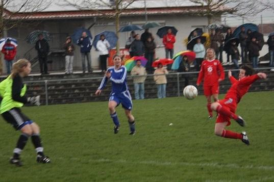 Szenenbild eines Fu&szlig;ballspiels bei Regen mit Spielerinnen in blauen und roten Trikots, Zuschauer mit Regenschirmen im Hintergrund.