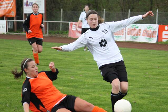 Zwei Fu&szlig;ballspielerinnen im Spiel: Eine in orange beim Rutschen, w&auml;hrend die andere in wei&szlig; den Ball spielt.
