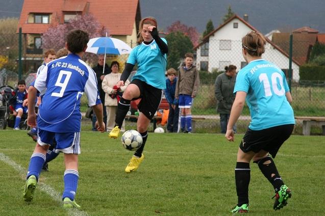 Fu&szlig;ballspiel auf einem Rasenplatz: Spieler in blauen und gr&uuml;nen Trikots k&auml;mpfen um den Ball, im Hintergrund Zuschauer.