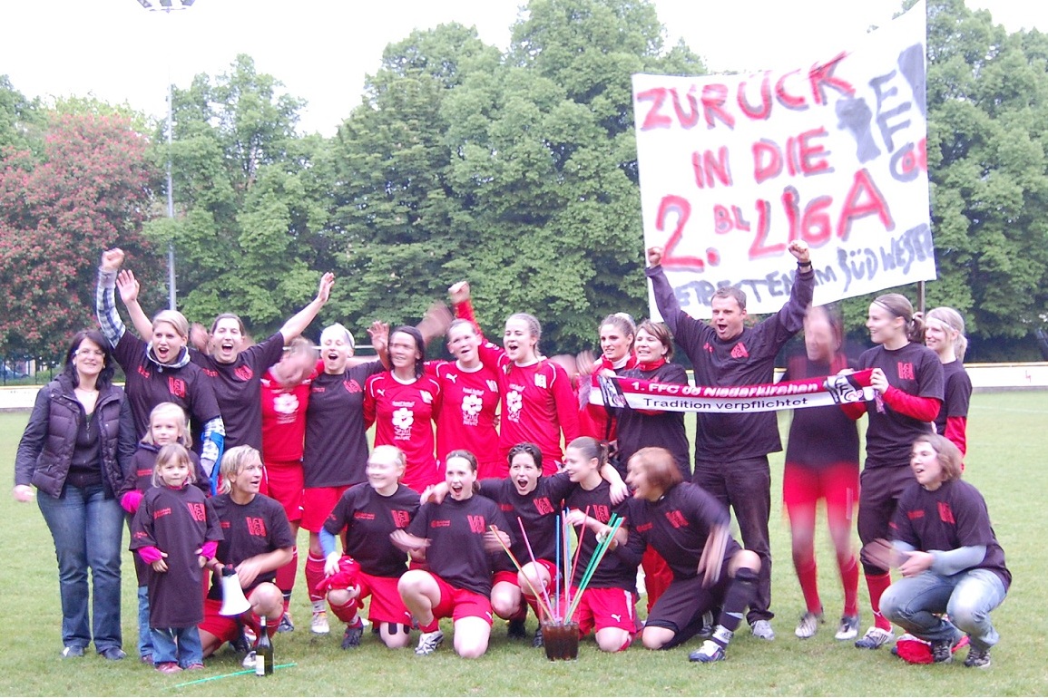 Gruppenszene von Fu&szlig;ballspielerinnen in roten Trikots, die jubeln, umgeben von Unterst&uuml;tzern mit einem Banner.
