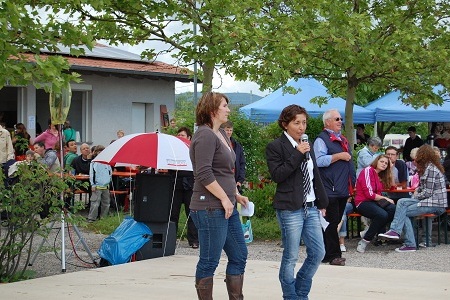 Zwei Frauen sprechen auf einer B&uuml;hne vor einer Menge, die an Tischen im Freien sitzt, unter B&auml;umen und Sonnenschirmen.