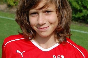 Porträt eines jungen Mädchens mit lockigem Haar in rotem Fußballtrikot auf einem grünen Fußballplatz.