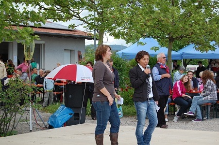 Zwei Frauen sprechen auf einer B&uuml;hne vor einer Menge, die an Tischen im Freien sitzt, unter B&auml;umen und Sonnenschirmen.