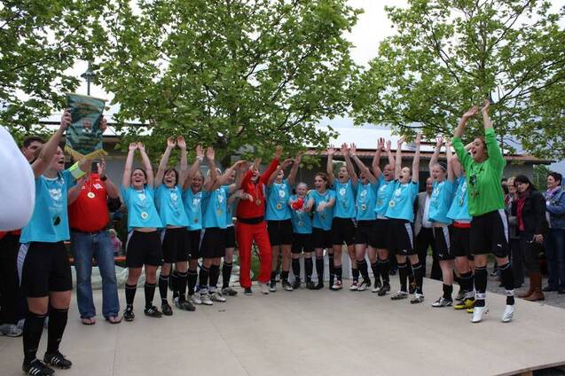 Feiernde Fu&szlig;ballmannschaft in blauen Trikots hebt Pokal und Medaillen, umgeben von jubelnden Fans und B&auml;umen.