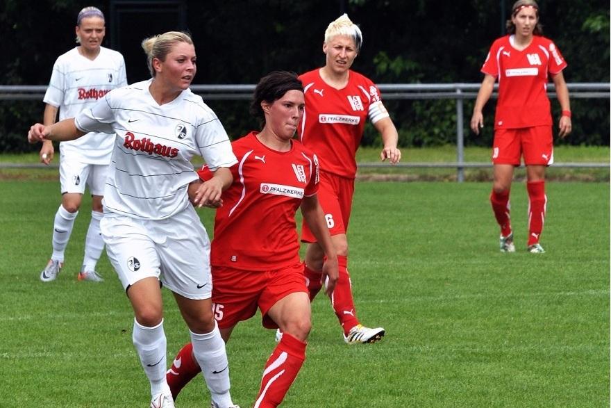 Zwei Fu&szlig;ballspielerinnen k&auml;mpfen um den Ball auf einem Fu&szlig;ballplatz, w&auml;hrend weitere Spielerinnen im Hintergrund positioniert sind.