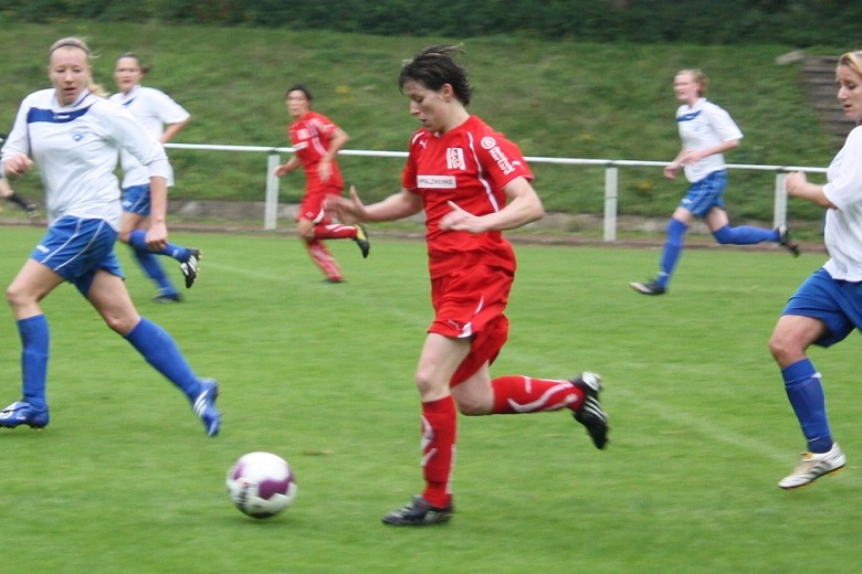 Spielszene eines Fu&szlig;ballspiels mit Spielerinnen in roten und blauen Trikots auf einem gr&uuml;nen Rasenplatz.