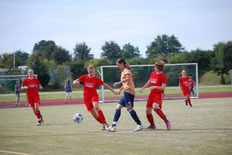 Zwei Fu&szlig;ballspielerinnen in roten Trikots und eine in gelbem Trikot k&auml;mpfen um den Ball auf einem Kunstrasenplatz.