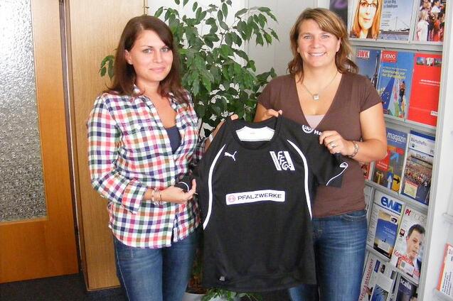 Zwei Frauen halten ein schwarzes Fu&szlig;balltrikot mit Logo und Sponsor an einem Pflanzenhintergrund in einem B&uuml;roumfeld.
