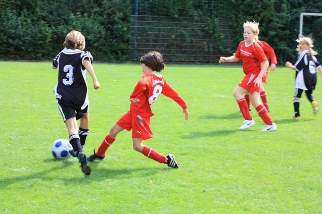 Zwei Kinder im Fu&szlig;balltrikot k&auml;mpfen um den Ball auf einem gr&uuml;nen Sportplatz. Im Hintergrund stehen weitere Spieler.