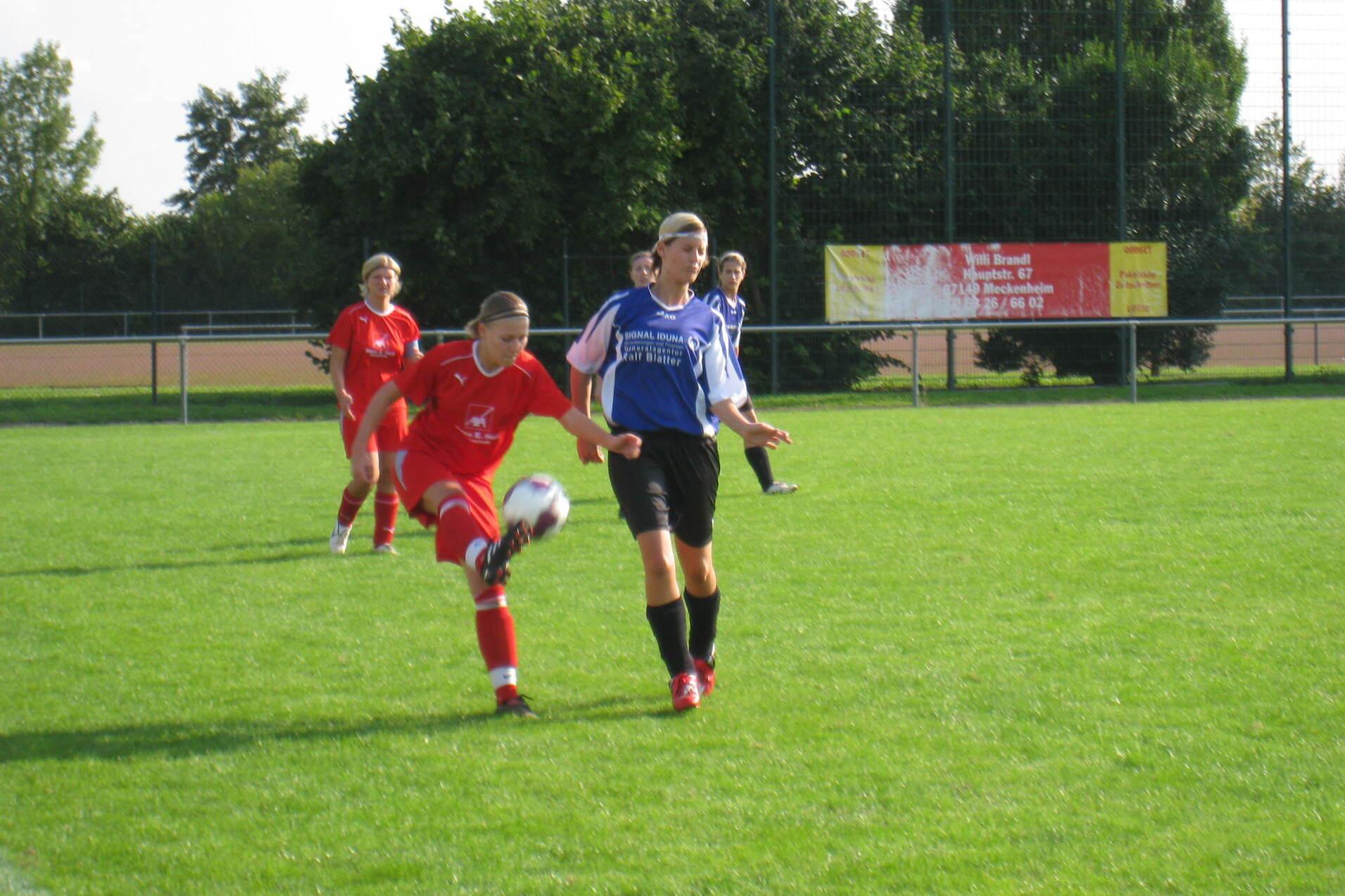 Zwei Fußballspielerinnen auf dem Feld, eine in rotem und eine in schwarzem Trikot, verfolgen einen Ball.