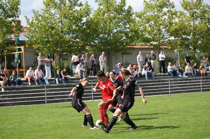 Zwei Spielerinnen in Schwarzem und ein Spieler in Rot k&auml;mpfen um den Ball auf einem Fu&szlig;ballplatz. Zuschauer sitzen im Hintergrund.