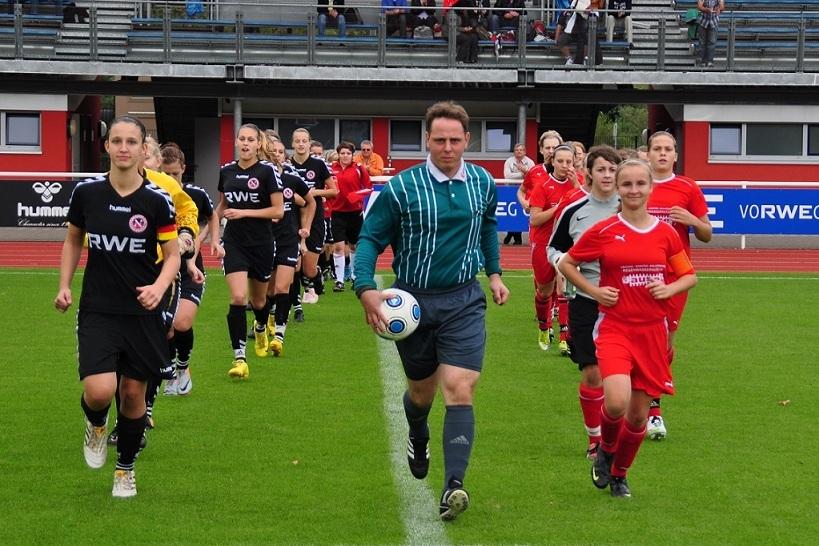 Fußballspielerinnen in schwarzen und roten Trikots betreten mit einem Schiedsrichter das Spielfeld.