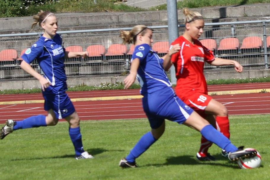 Zwei Fu&szlig;ballspielerinnen in blauen Trikots verfolgen eine Spielerin in rotem Trikot auf einem Rasenplatz.
