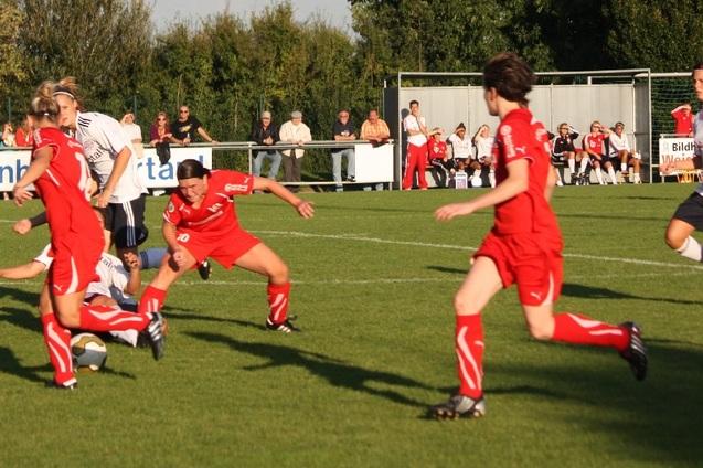 Szenenbild eines Fu&szlig;ballspiels, bei dem Spielerinnen in roten Trikots um den Ball k&auml;mpfen, w&auml;hrend Zuschauer im Hintergrund stehen.