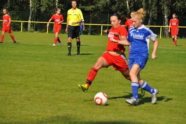 Zwei Fu&szlig;ballspielerinnen in roten und blauen Trikots k&auml;mpfen um den Ball auf einem gr&uuml;nen Spielfeld.