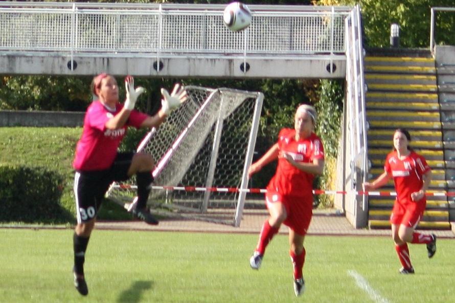 Torhüterin in pink springt nach einem Ball, während zwei Spielerinnen in rot auf dem Feld agieren.