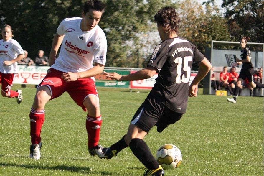 Zwei Fu&szlig;ballspieler im Wettkampf, einer mit wei&szlig;em Trikot und roter Hose, der andere im schwarzen Trikot.