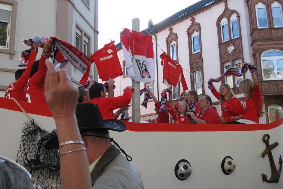 Gruppen von Menschen in roten T-Shirts und Schals feiern auf einem festlichen Wagen vor historischen Geb&auml;uden.