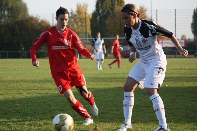 Zwei Fußballspielerinnen im Wettkampf um den Ball auf einem grünen Spielfeld, umgeben von herbstlichen Bäumen.