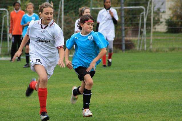 Zwei Kinder rennen auf einem Fu&szlig;ballfeld, eines tr&auml;gt ein rotes und wei&szlig;es Trikot, das andere ein blaues Trikot.