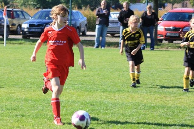 Junge in rotem Fu&szlig;balltrikot dribbelt den Ball auf einem Rasenplatz mit Zuschauern im Hintergrund.