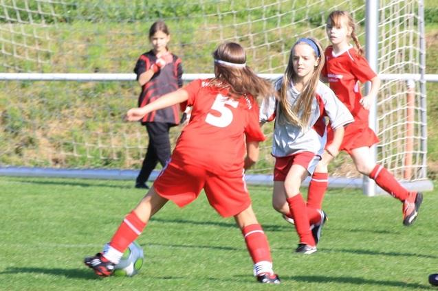 Vier M&auml;dchen im Fu&szlig;balltrikot spielen auf einem gr&uuml;nen Feld, w&auml;hrend eine Spielerin den Ball dribbelt.