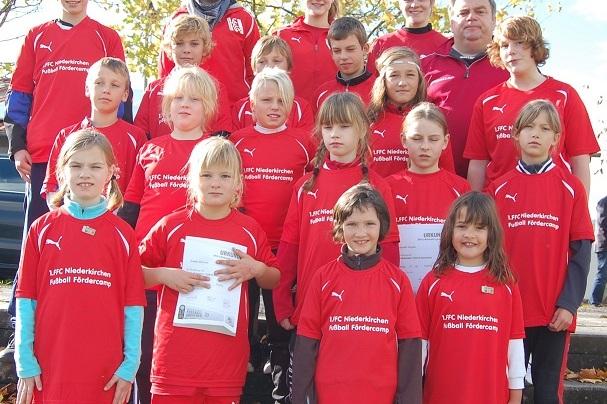 Gruppe von Kindern in roten Fu&szlig;balltrikots mit Betreuern, alle stehen im Freien und l&auml;cheln in die Kamera.