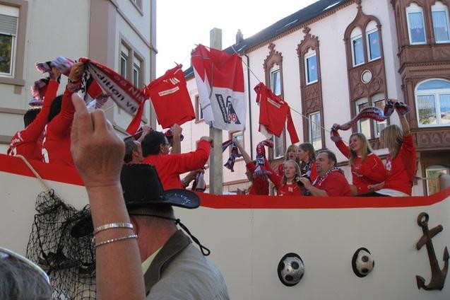 Gruppen von Menschen in roten T-Shirts und Schals feiern auf einem festlichen Wagen vor historischen Geb&auml;uden.