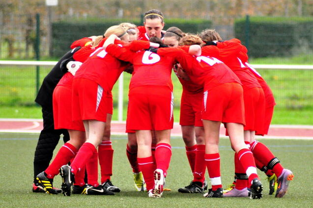 Mannschaftsbesprechung einer Fu&szlig;ballmannschaft in roten Trikots, Spielerinnen umarmen sich auf dem Spielfeld.
