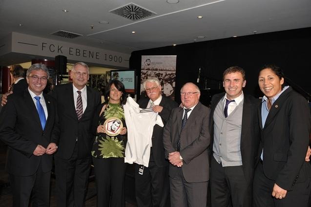Sechs Personen stehen l&auml;chelnd zusammen, eine h&auml;lt ein Trikot, im Hintergrund eine B&uuml;hne und eine Tafel.