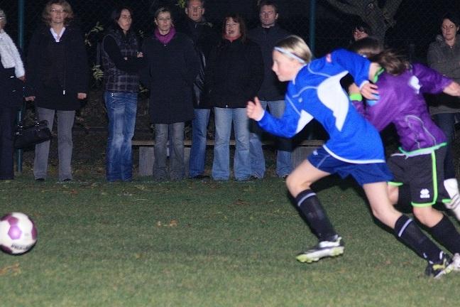 Zwei Mädchen in Sportkleidung jagen einem Fußball auf einem Platz hinter zuschauenden Personen.