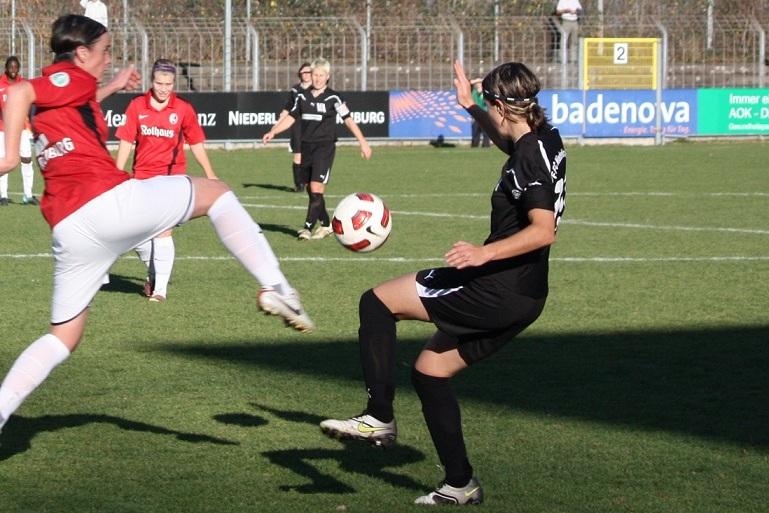 Zwei Fu&szlig;ballspielerinnen im Wettkampf, eine in rotem, die andere in schwarzem Trikot, w&auml;hrend der Ball in der Luft ist.