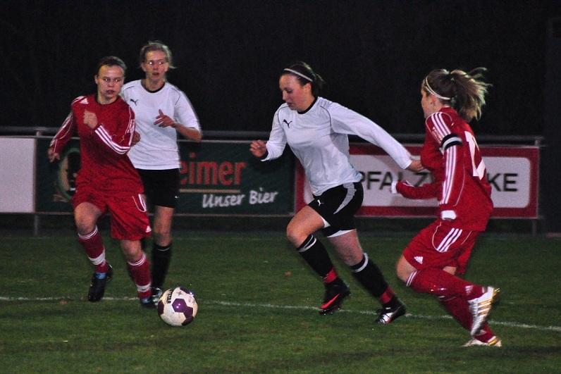 Zwei Fußballspielerinnen in roten Trikots verfolgen eine Spielerin in einem weißen Trikot auf einem Kunstrasenplatz.