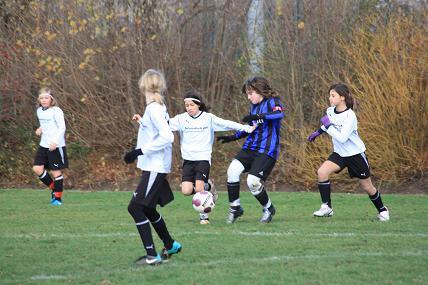 M&auml;dchenfu&szlig;ballspiel auf einem Rasenplatz, mehrere Spielerinnen in wei&szlig;en und gestreiften Trikots verfolgen den Ball.