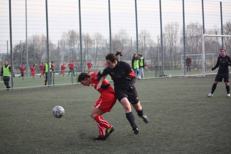 Zwei Frauen im Fu&szlig;ball, eine in Rot und eine in Schwarz, k&auml;mpfen um den Ball auf einem Kunstrasenplatz.