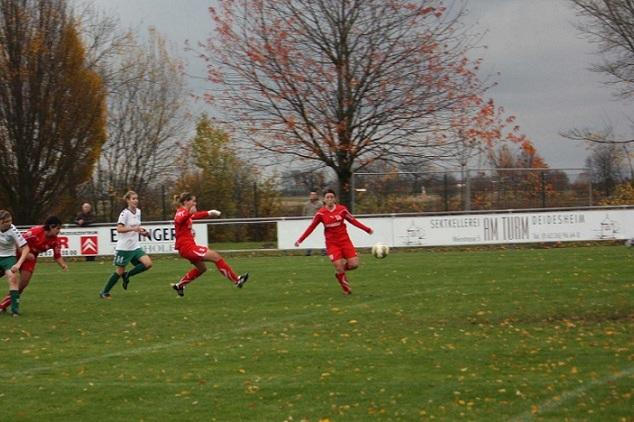 Zwei Fu&szlig;ballspielerinnen in Rot dribbeln auf einem Rasenplatz, w&auml;hrend eine Torh&uuml;terin in Blau zum Schuss bereit steht.