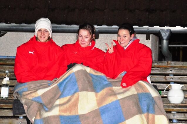 Drei Frauen in roten Jacken sitzen auf einer Trib&uuml;ne, zugedeckt mit einer Wolldecke, und l&auml;cheln in die Kamera.