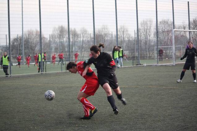 Zwei Frauen im Fu&szlig;ball, eine in Rot und eine in Schwarz, k&auml;mpfen um den Ball auf einem Kunstrasenplatz.