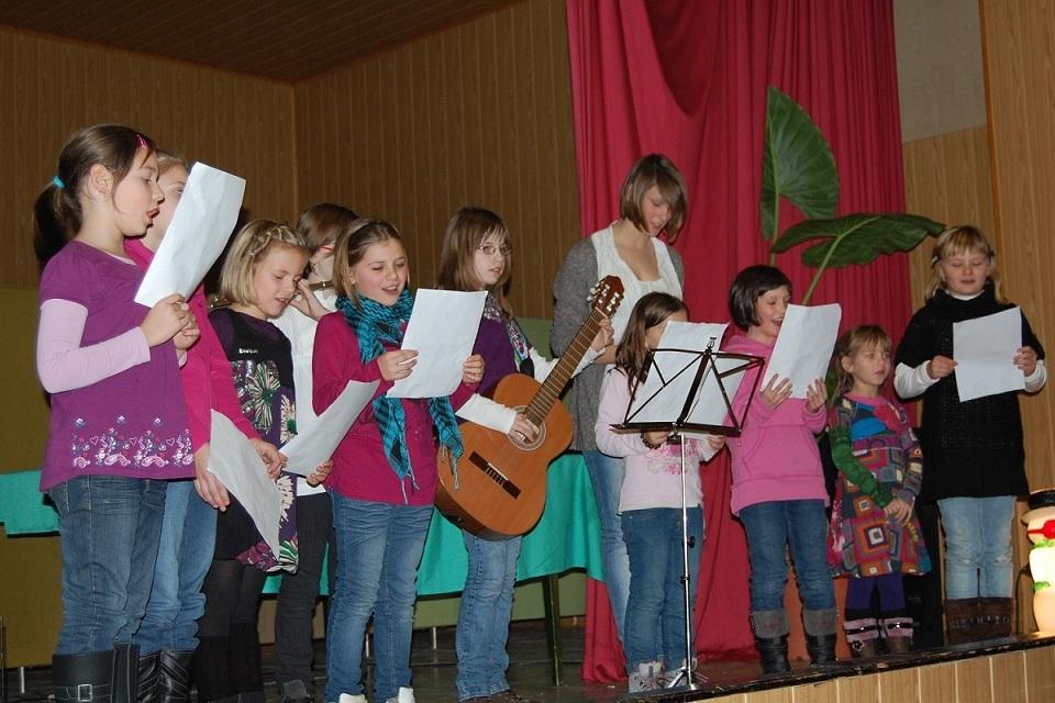 Mädchen stehen auf einer Bühne und singen mit Textblättern, während eine von ihnen eine Gitarre spielt.