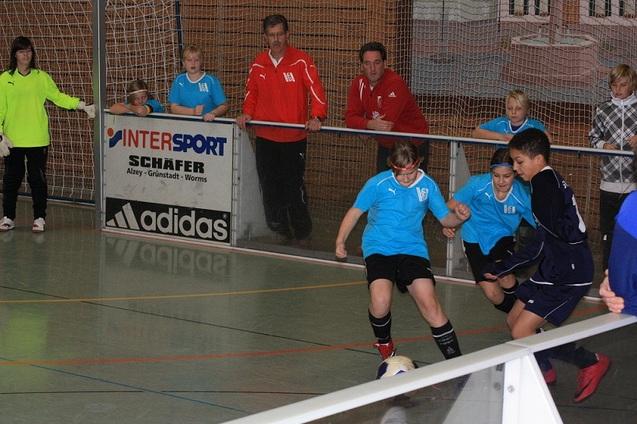 Zwei Kinder spielen Fu&szlig;ball in einer Halle, w&auml;hrend Trainer und Zuschauer am Rand beobachten.