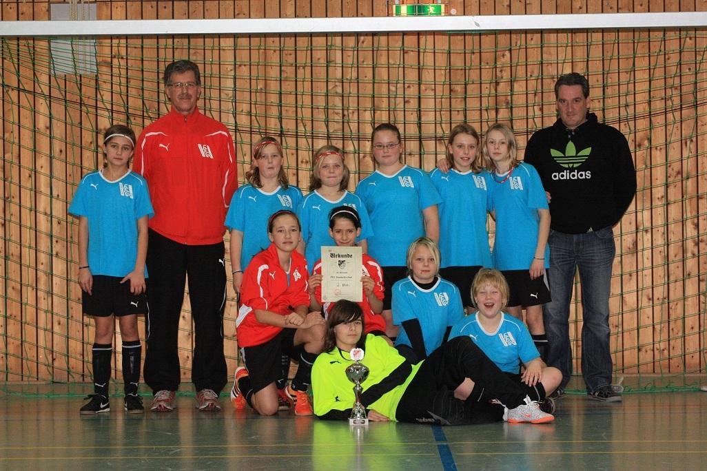 Mannschaftsbild einer M&auml;dchenfu&szlig;ballmannschaft mit Pokal und Urkunde in einer Sporthalle, umgeben von Trainern.