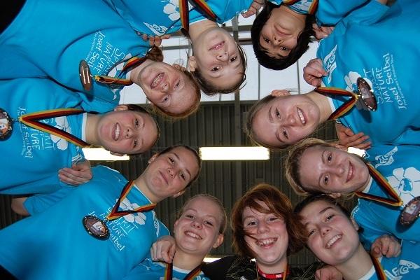 Gruppe von 10 M&auml;dchen in blauen T-Shirts mit Medaillen, die in einem Kreis l&auml;cheln und in die Kamera schauen.