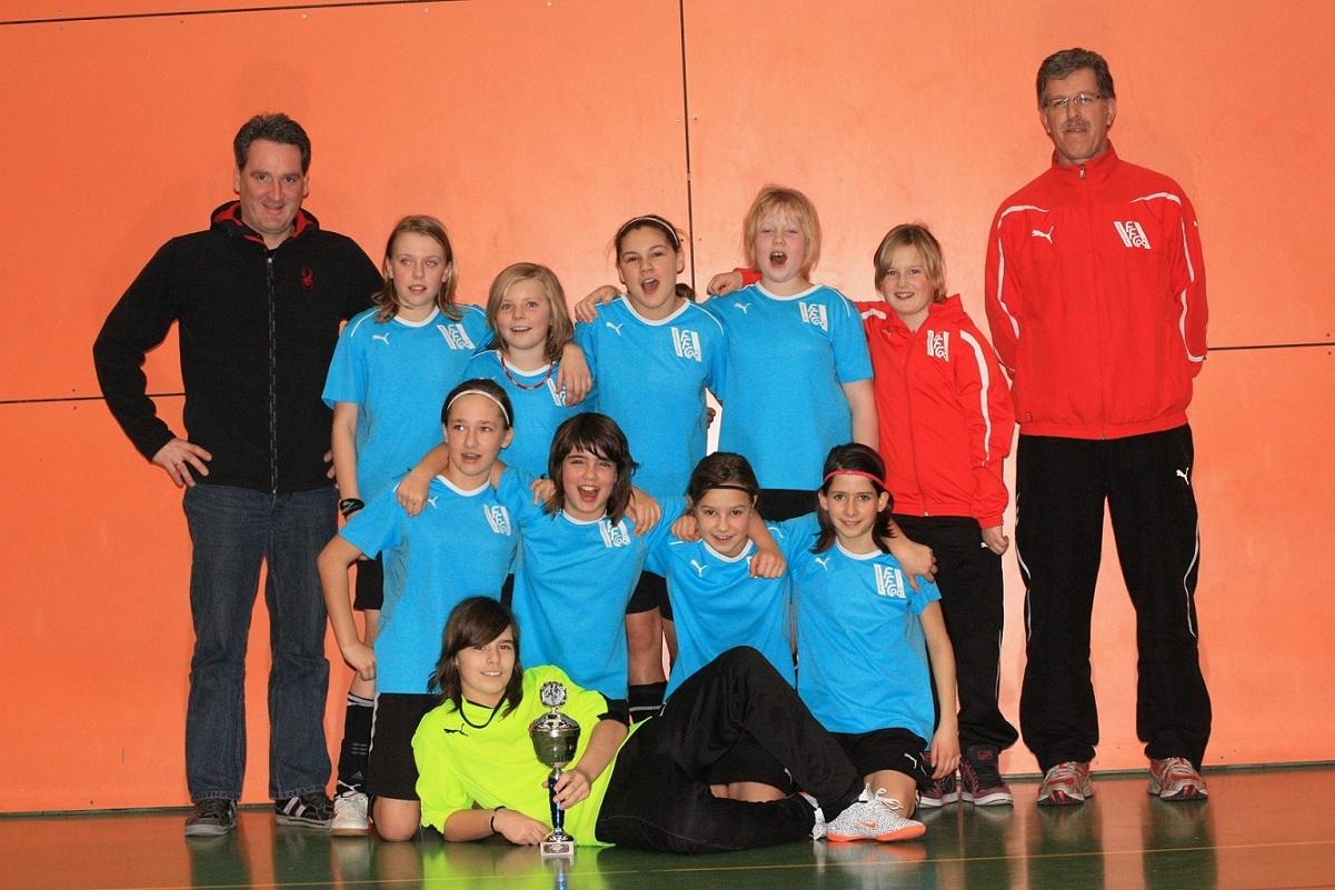 Kinderfu&szlig;ballmannschaft mit Pokal, aufgeteilt in zwei Reihen, vor einer orangefarbenen Wand, Trainer stehen hinter ihnen.