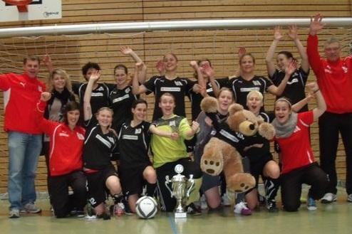 Feiernde Frauenmannschaft im Hallenfu&szlig;ball mit Pokal, Ball und einem Stoffb&auml;ren in sportlicher Kleidung.