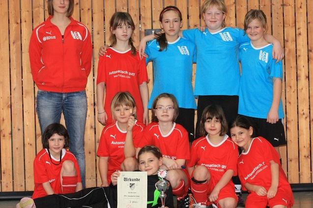 Gruppensfoto einer M&auml;dchenfu&szlig;ballmannschaft mit Trainerin, Pokal und Urkunde in einer Sporthalle.