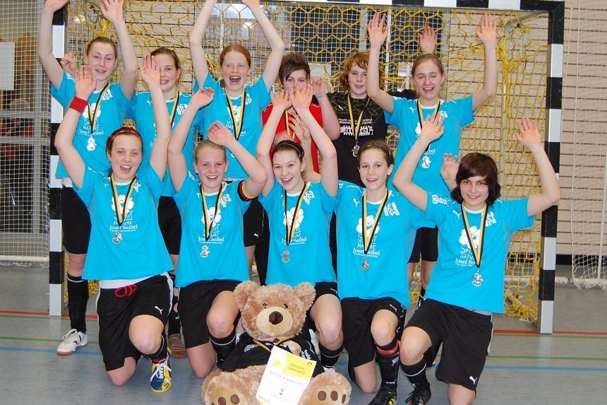M&auml;dchen-Handballteam feiert mit erhobenen H&auml;nden, Silbermedaillen und einem Teddyb&auml;r vor einem Tor.