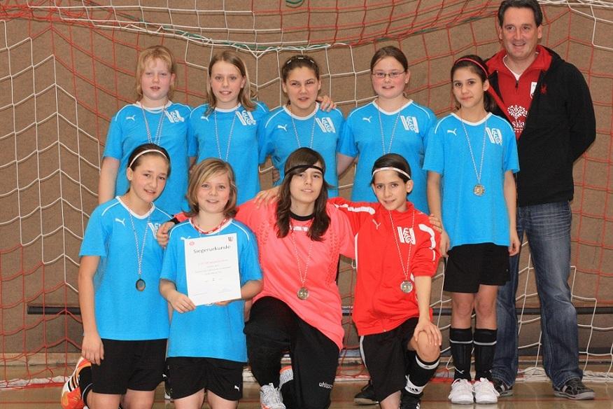 M&auml;dchenfu&szlig;ballmannschaft in blauen Trikots mit Medaillen posiert vor einem Fu&szlig;balltor, ein M&auml;dchen h&auml;lt eine Urkunde.