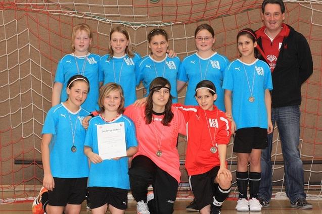 M&auml;dchenfu&szlig;ballmannschaft in blauen Trikots mit Medaillen posiert vor einem Fu&szlig;balltor, ein M&auml;dchen h&auml;lt eine Urkunde.